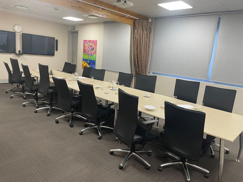 A functional office boardroom featuring a long conference table, carpeted flooring, recessed ceiling lighting, and roller blinds for privacy.