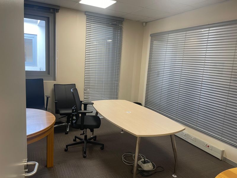 A functional office space featuring carpeted flooring, recessed ceiling lighting, and large windows fitted with adjustable venetian blinds.