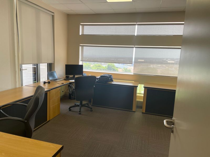 A professional office space featuring built-in wooden desks, carpeted flooring, and large windows with roller blinds providing natural light and exterior views.