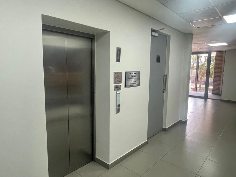 A clean office building hallway featuring a stainless steel elevator, a secure access door, and modern tiled flooring with recessed ceiling lighting.