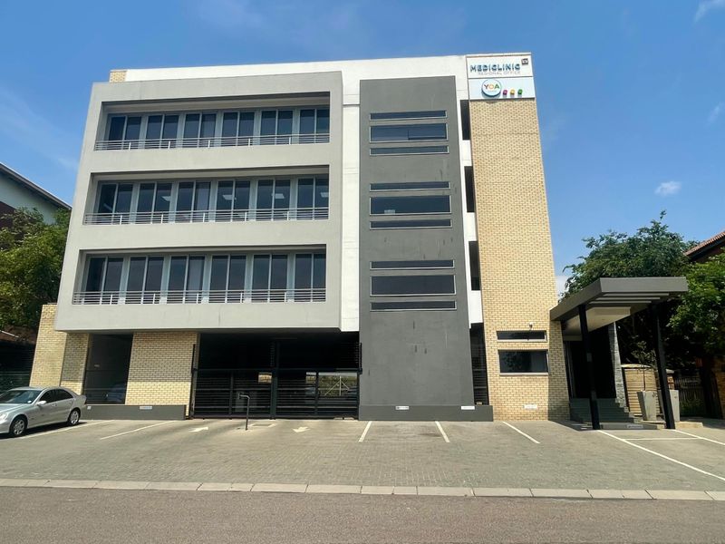 A modern multi-story office building featuring a brick and concrete facade, large windows, and a paved parking area at the front entrance.