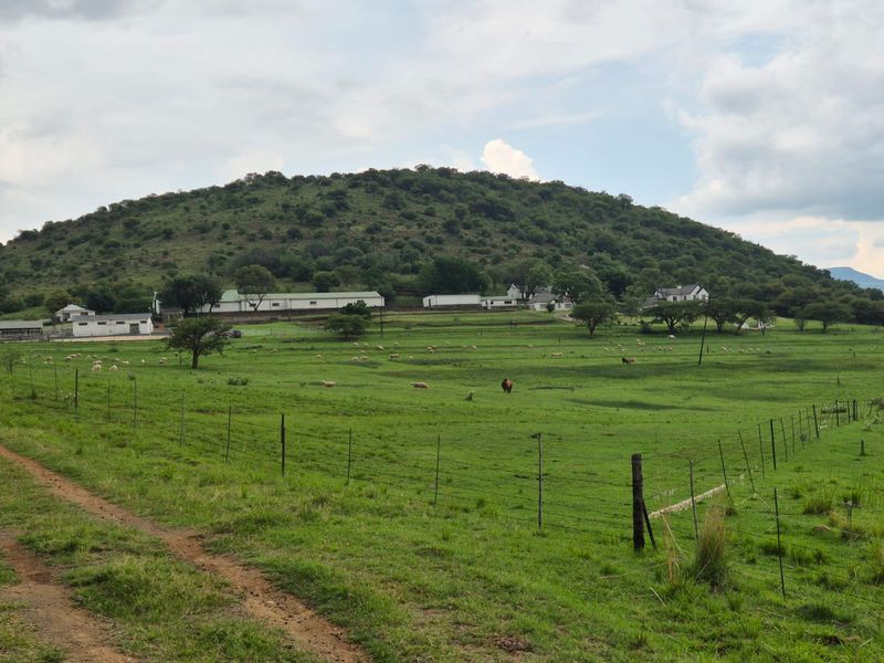 A scenic view of a rural farm property featuring expansive green pastures, wire fencing, and a collection of farm buildings set against a hill.