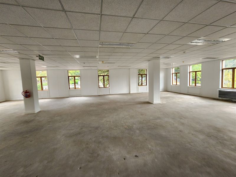 A spacious, open-plan office area featuring a suspended ceiling, recessed lighting, structural columns, and large windows providing ample natural light.