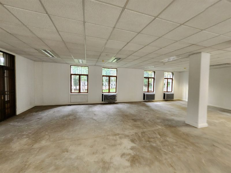 Spacious, open-plan office area featuring a suspended ceiling, recessed lighting, window-mounted air conditioning units, and large windows with security bars.