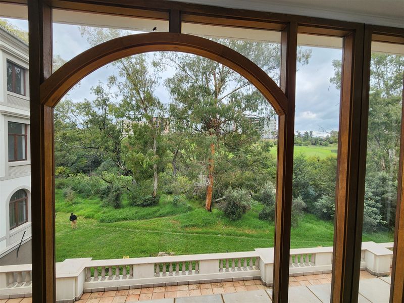 A view from an interior space looking out through a timber-framed arched window onto a green garden area and balcony with a stone balustrade.