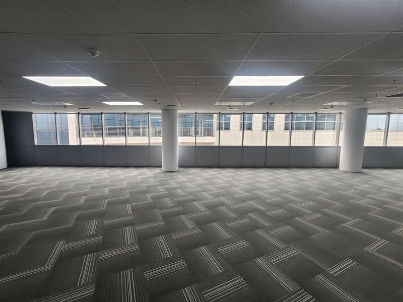 A spacious, empty open-plan office area featuring carpet tiles, suspended ceilings with recessed lighting, and large windows providing natural light.