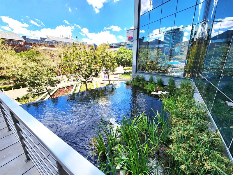 Modern office building exterior featuring a reflective glass facade, a tranquil water feature, and landscaped garden area with metal safety railings.