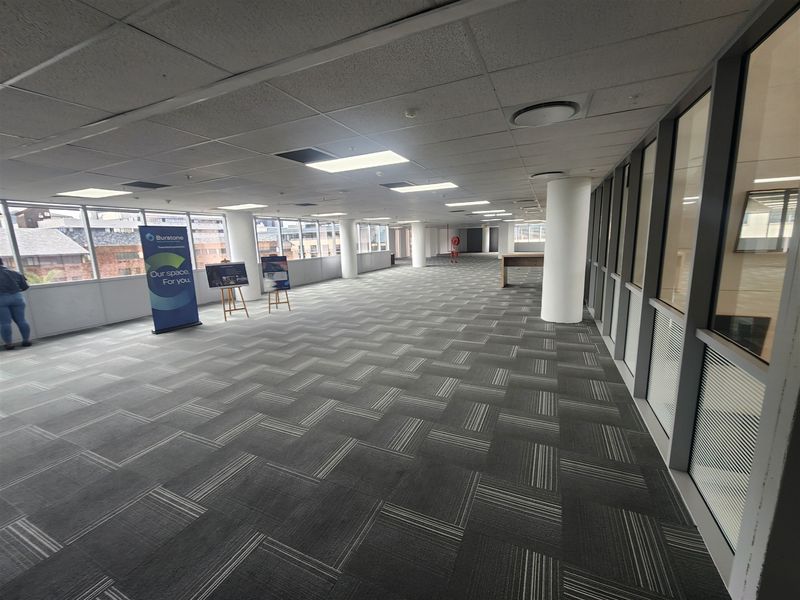 Spacious open-plan office area featuring carpet tiles, structural columns, and extensive glass windows providing natural light and views.