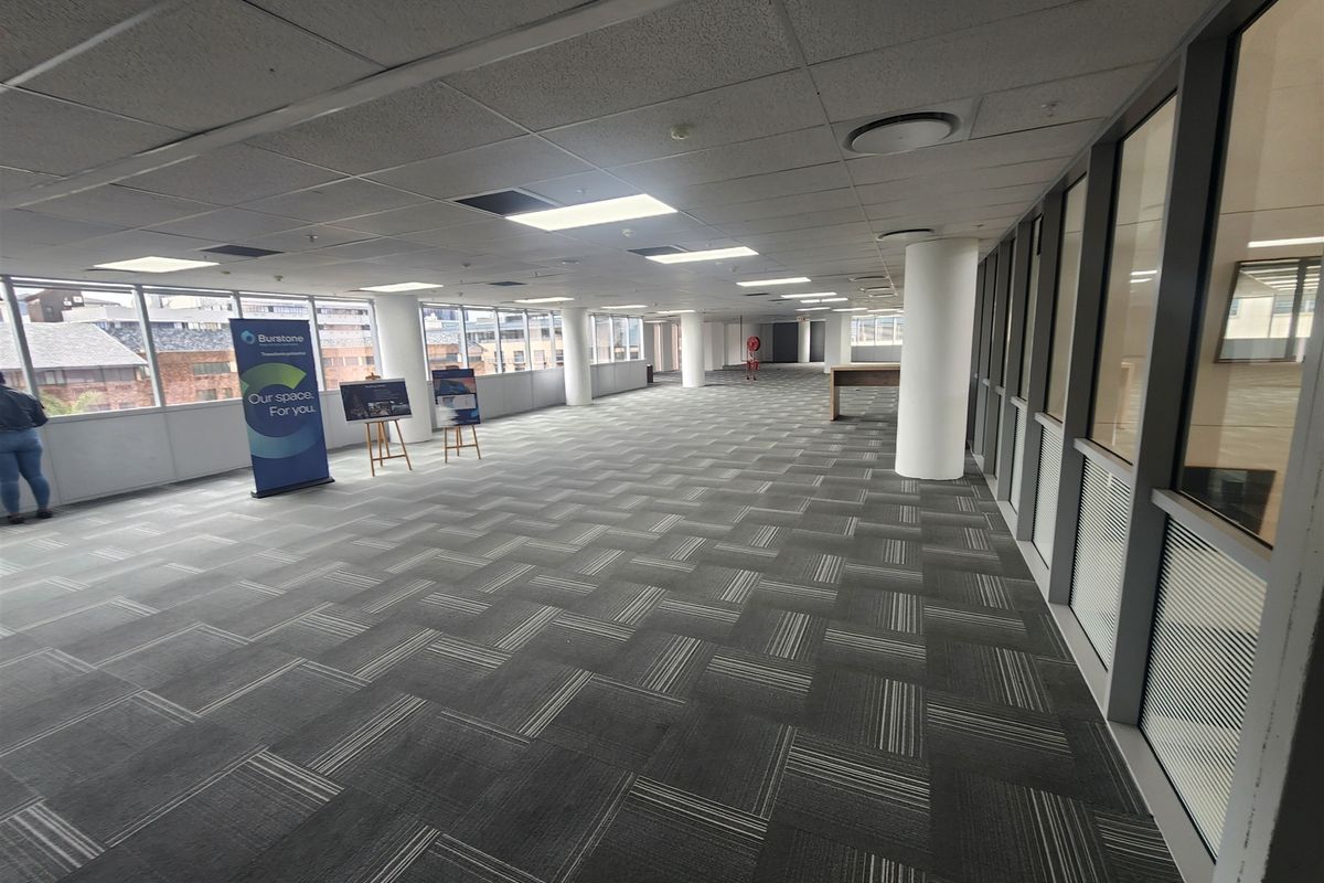 Spacious open-plan office area featuring carpet tiles, structural columns, and extensive glass windows providing natural light and views.