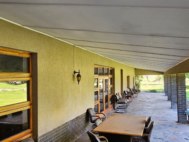 A spacious covered patio featuring stone flooring, brick support pillars, and large wooden-framed windows overlooking the surrounding farm landscape.