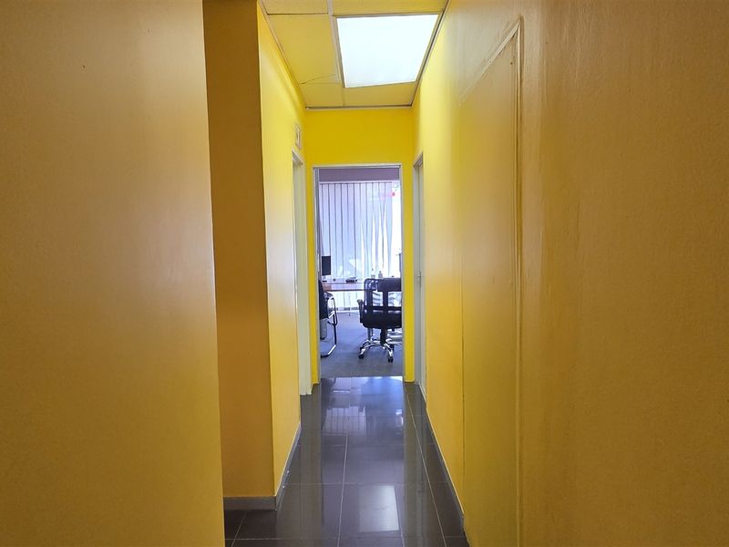 A narrow office hallway featuring tiled flooring, bright yellow walls, and a drop ceiling with recessed lighting, leading into an adjacent office space.