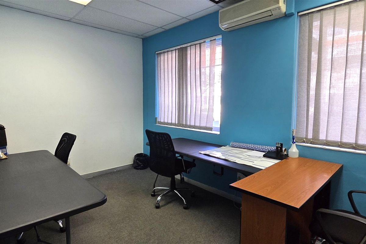 A functional office space featuring carpeted flooring, a suspended ceiling, split-system air conditioning, and large windows with vertical blinds.