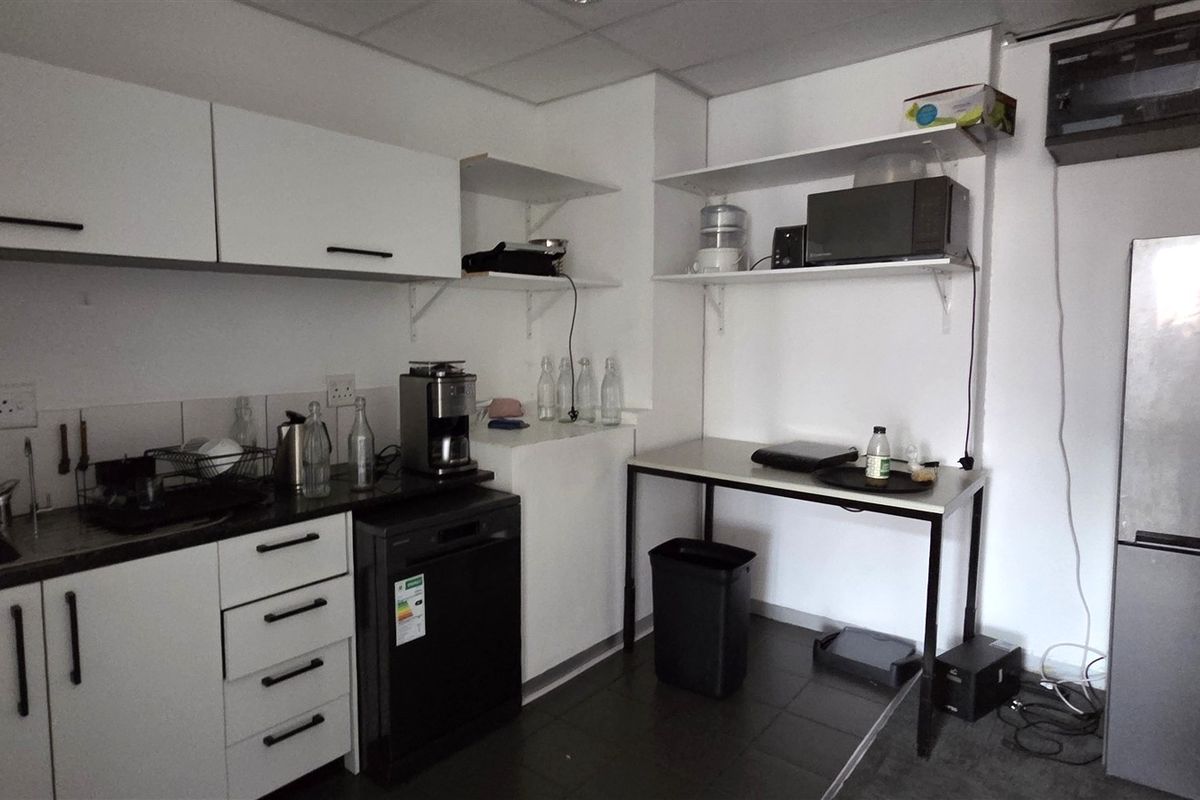 A functional office kitchenette featuring white cabinetry, stainless steel sink, open shelving, and tiled flooring, providing a practical space for staff refreshments.