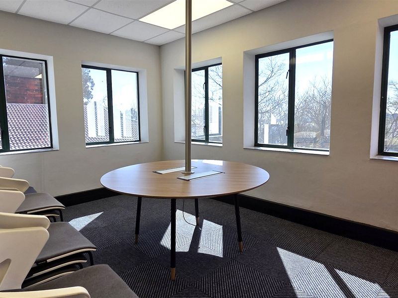 A bright office space featuring large windows with natural light, carpeted flooring, and a central meeting table.