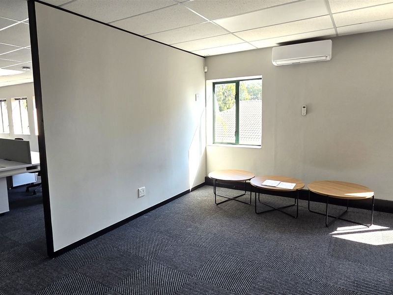 A bright, open-plan office space featuring carpeted flooring, a suspended ceiling, and a split-system air conditioning unit, providing a functional professional work environment.