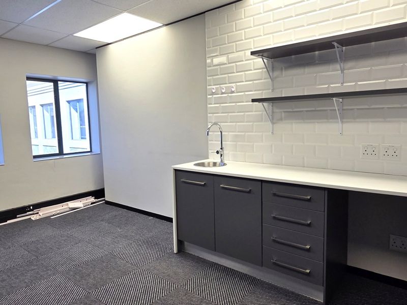 Modern office space featuring a built-in kitchenette with subway tile backsplash, floating shelves, sink, carpet tiles, and ample natural light from large windows.