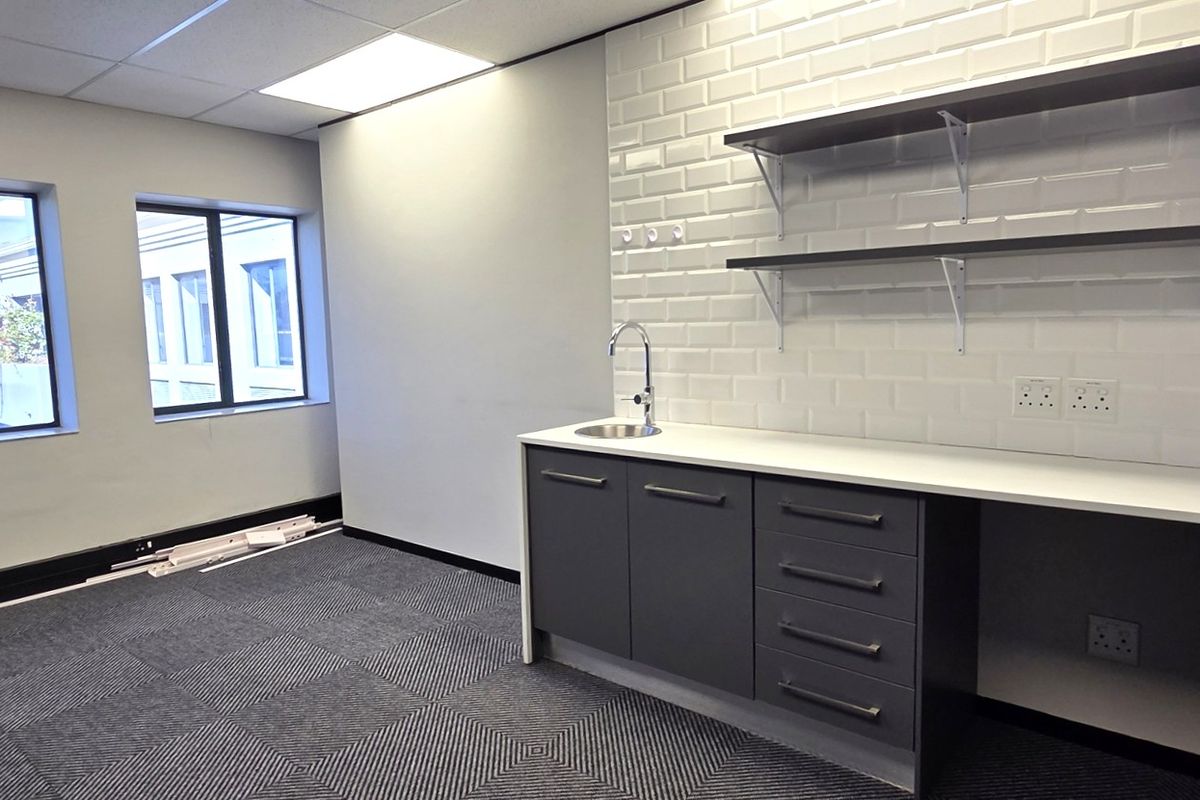 Modern office space featuring a built-in kitchenette with subway tile backsplash, floating shelves, sink, carpet tiles, and ample natural light from large windows.