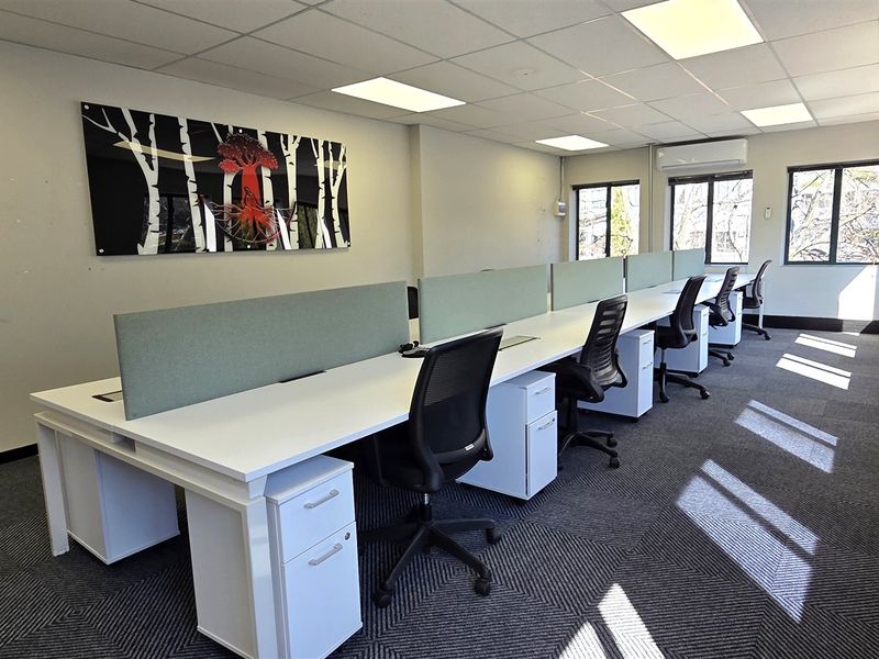 A bright, modern office space featuring a long workstation configuration, suspended ceiling with recessed lighting, air conditioning, and large windows providing natural light.