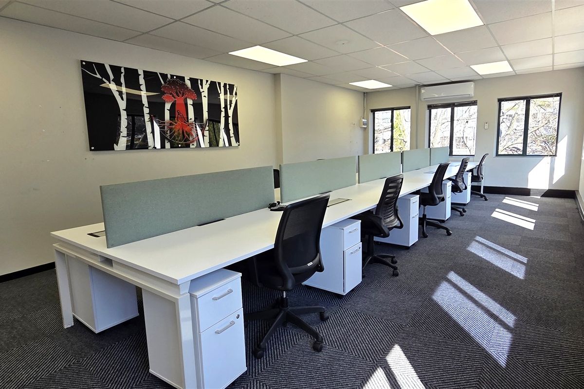 A bright, modern office space featuring a long workstation configuration, suspended ceiling with recessed lighting, air conditioning, and large windows providing natural light.