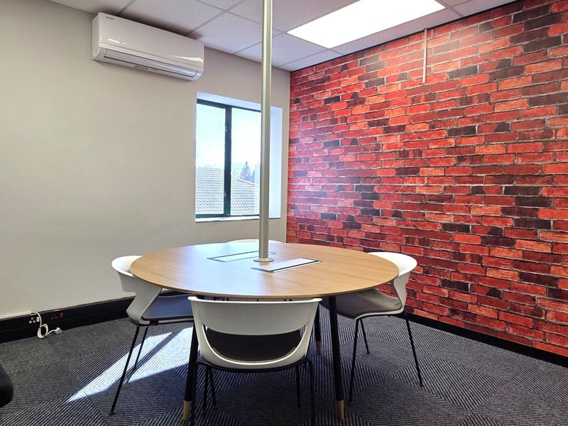 A modern office space featuring a central meeting table, carpeted flooring, an exposed brick feature wall, and a wall-mounted air conditioning unit.