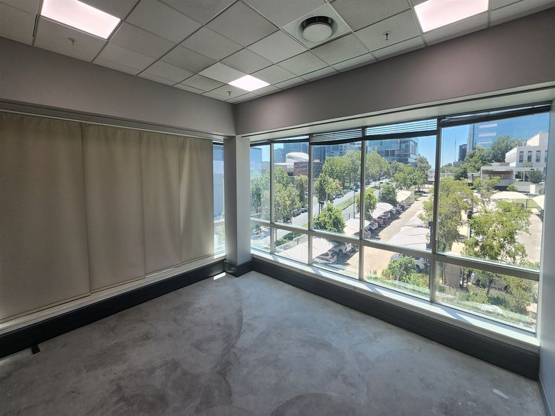 A bright, empty office space featuring large windows with views, suspended ceilings with recessed lighting, and unfinished concrete flooring.