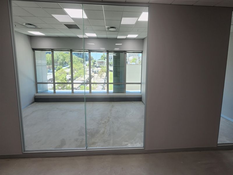 A bright, empty office space featuring a glass partition wall, suspended ceiling with recessed lighting, and large windows offering an urban view.