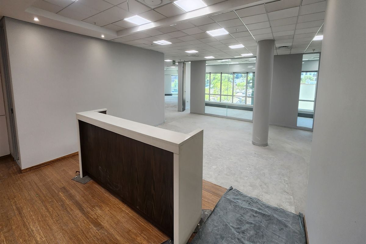An open-plan office space featuring a built-in reception desk, suspended ceiling with recessed lighting, and large windows providing natural light.