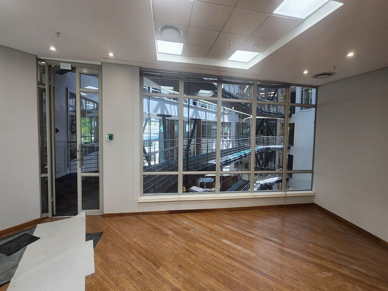 A bright, empty office space featuring wooden flooring, recessed ceiling lighting, and a large glass wall overlooking an internal atrium.