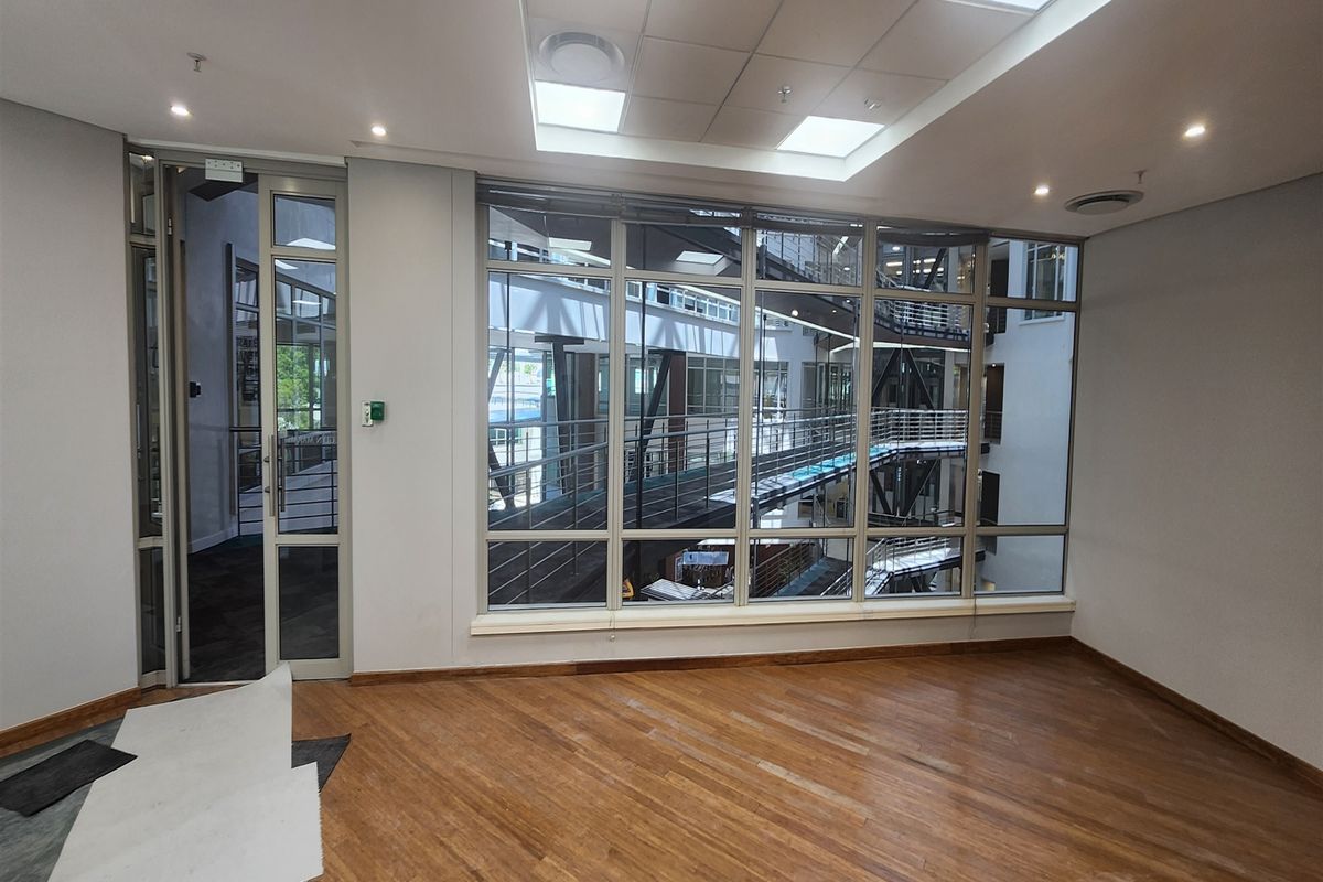 A bright, empty office space featuring wooden flooring, recessed ceiling lighting, and a large glass wall overlooking an internal atrium.