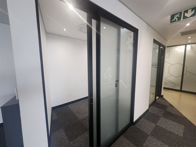A modern, empty office space featuring a frosted glass sliding door, carpeted flooring, and recessed lighting within a professional commercial environment.