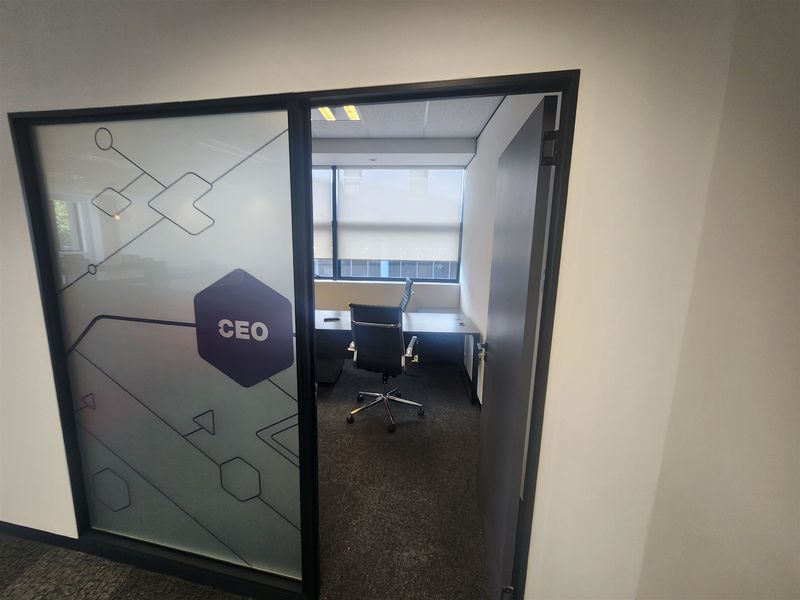A private office space featuring a frosted glass partition with branding, dark carpeting, and large windows with fitted blinds.