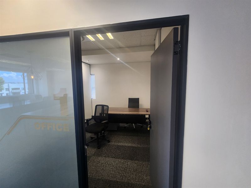 A professional office space featuring a glass partition entrance, recessed ceiling lighting, and dark carpet flooring.