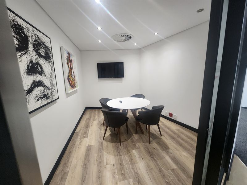 A modern, well-lit office meeting room featuring wood-look flooring, recessed ceiling lighting, and a wall-mounted television.