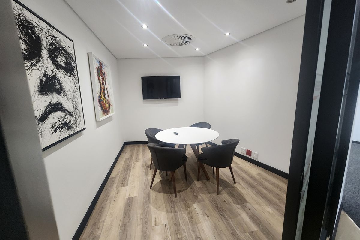A modern, well-lit office meeting room featuring wood-look flooring, recessed ceiling lighting, and a wall-mounted television.