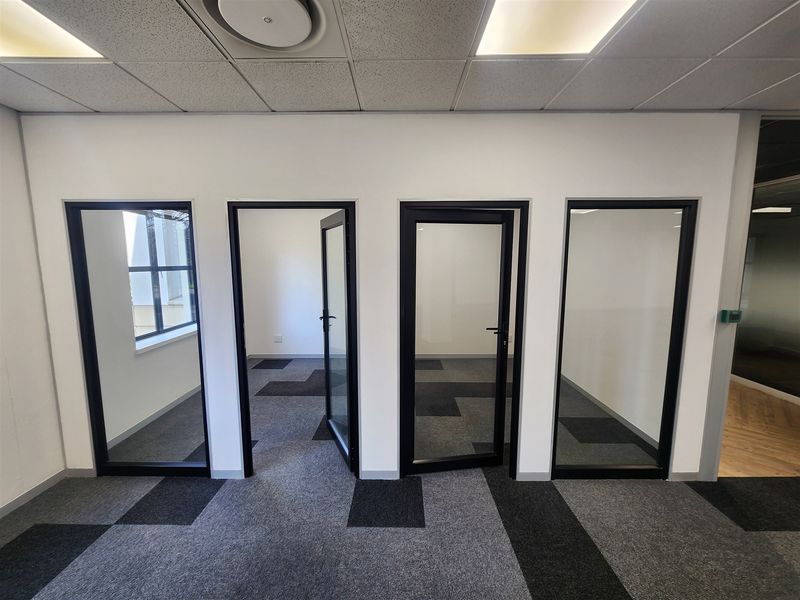 Modern office space featuring glass partitions, black aluminium frames, carpet tiles, and a suspended ceiling with recessed lighting.