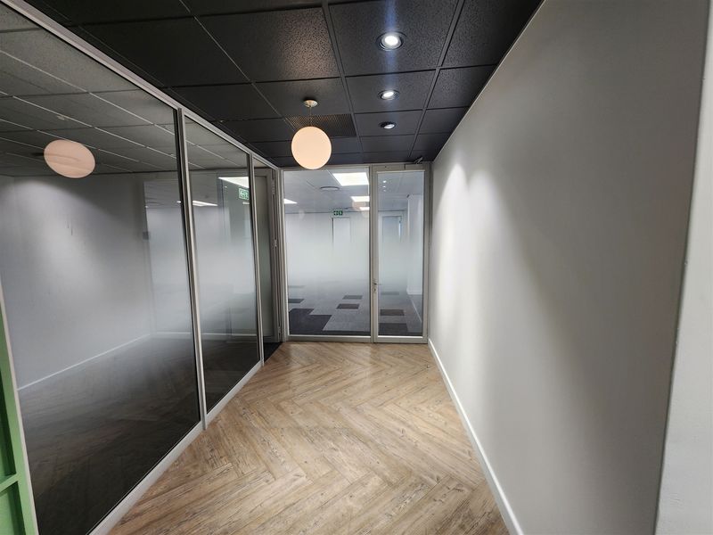 A modern office hallway featuring herringbone flooring, a dark suspended ceiling with recessed lighting, and glass-partitioned office spaces with frosted privacy film.