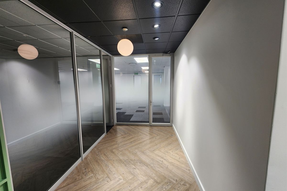 A modern office hallway featuring herringbone flooring, a dark suspended ceiling with recessed lighting, and glass-partitioned office spaces with frosted privacy film.