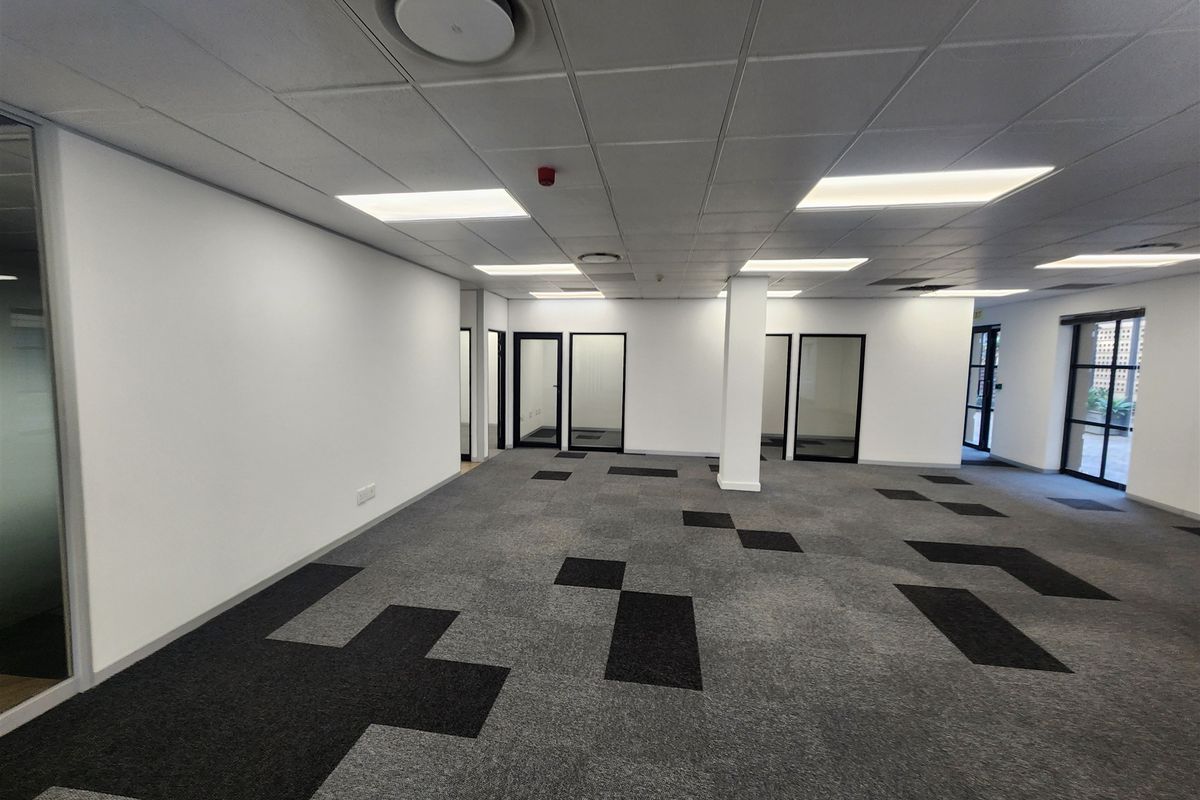 A spacious, open-plan office area featuring a suspended ceiling, recessed lighting, and modern patterned carpet tiles with glass-partitioned doorways.