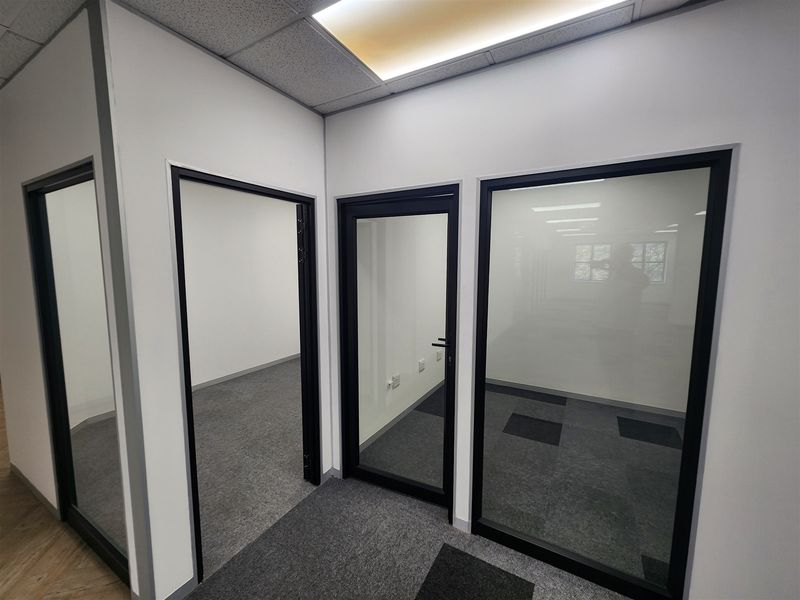 A modern, vacant office space featuring glass partitions, black aluminium frames, carpet tiles, and a suspended ceiling with recessed lighting.