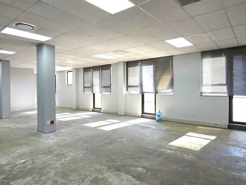 An open-plan office space featuring a suspended ceiling, recessed lighting, structural columns, and large windows with venetian blinds, currently awaiting floor finishing.