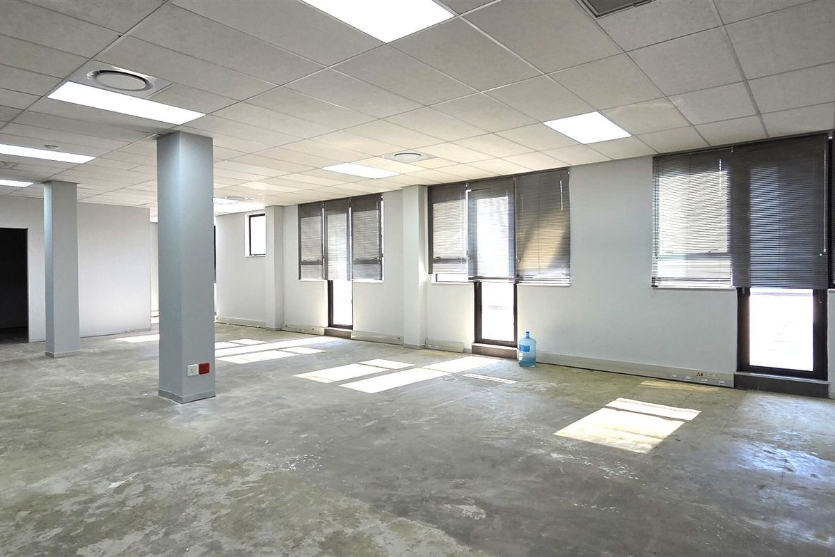 An open-plan office space featuring a suspended ceiling, recessed lighting, structural columns, and large windows with venetian blinds, currently awaiting floor finishing.