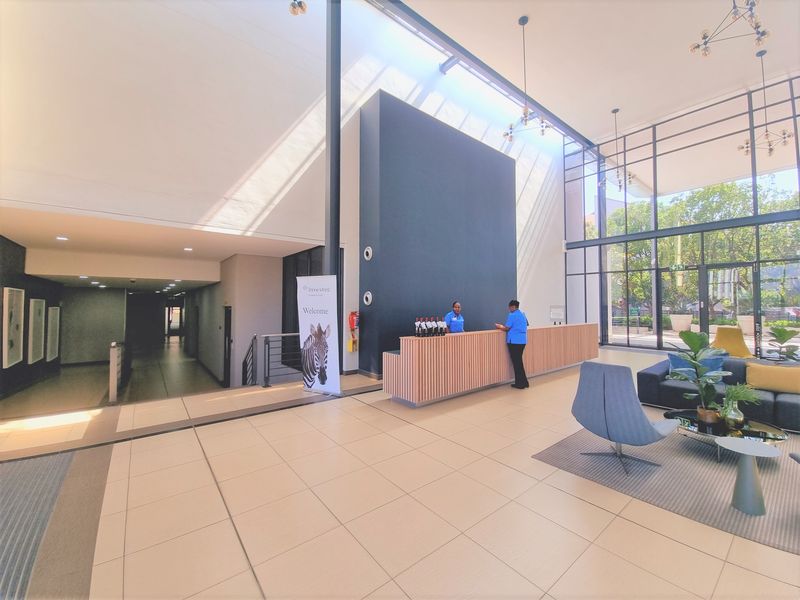 Spacious modern office reception area featuring high ceilings, floor-to-ceiling glass windows, tiled flooring, and a contemporary wooden reception desk.