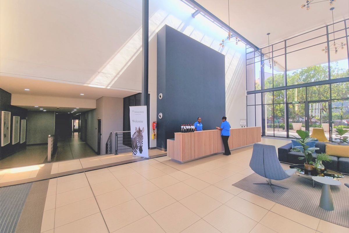 Spacious modern office reception area featuring high ceilings, floor-to-ceiling glass windows, tiled flooring, and a contemporary wooden reception desk.
