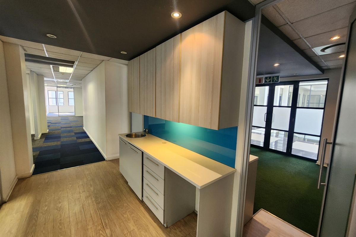 A modern office kitchenette featuring built-in cabinetry, a laminate countertop with a stainless steel sink, and a vibrant blue glass splashback.