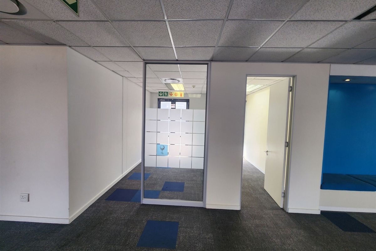 A vacant office space featuring carpet tiles, a suspended ceiling with recessed lighting, a glass partition, and a built-in blue bench seating area.