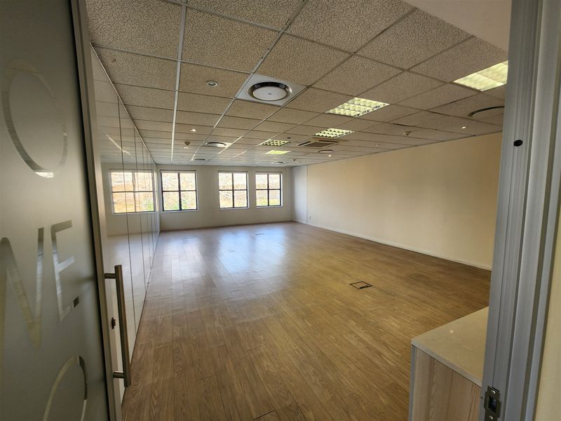 A spacious, vacant office area featuring wood-look flooring, a suspended ceiling with recessed lighting, and a modern glass partition wall.