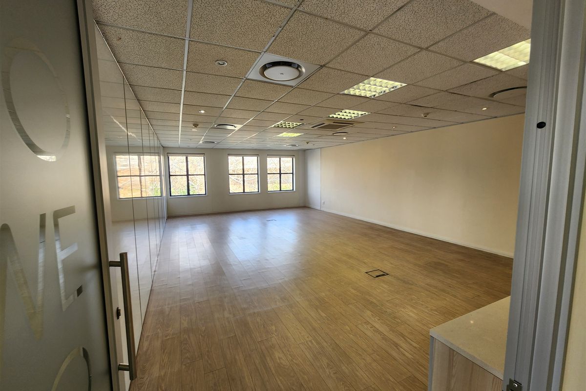 A spacious, vacant office area featuring wood-look flooring, a suspended ceiling with recessed lighting, and a modern glass partition wall.