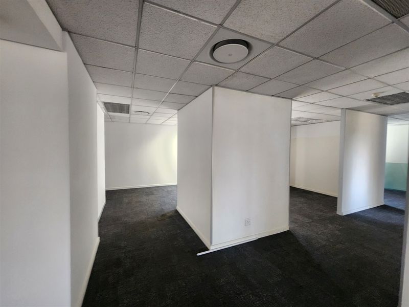 An open-plan office space featuring a suspended ceiling, recessed lighting, air conditioning vents, and dark carpeting throughout the vacant interior.