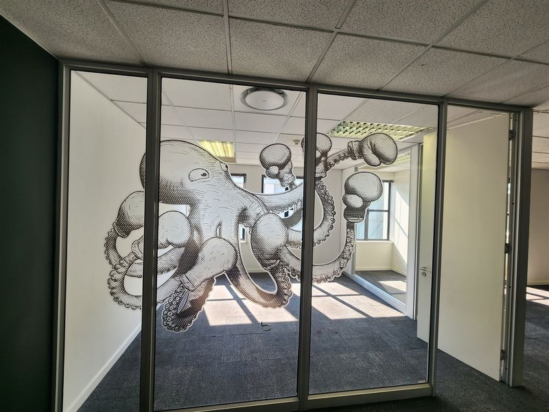 An empty office space featuring a glass partition wall with a decorative octopus graphic, suspended ceiling, and carpeted flooring.