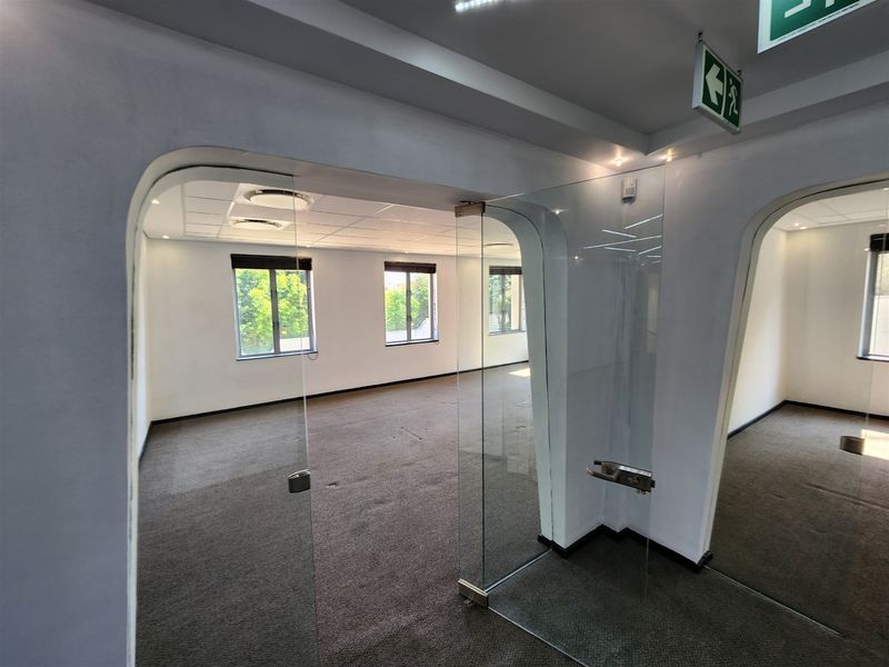 A bright, open-plan office space featuring modern glass partition doors, suspended ceilings with recessed lighting, and neutral carpeting throughout.